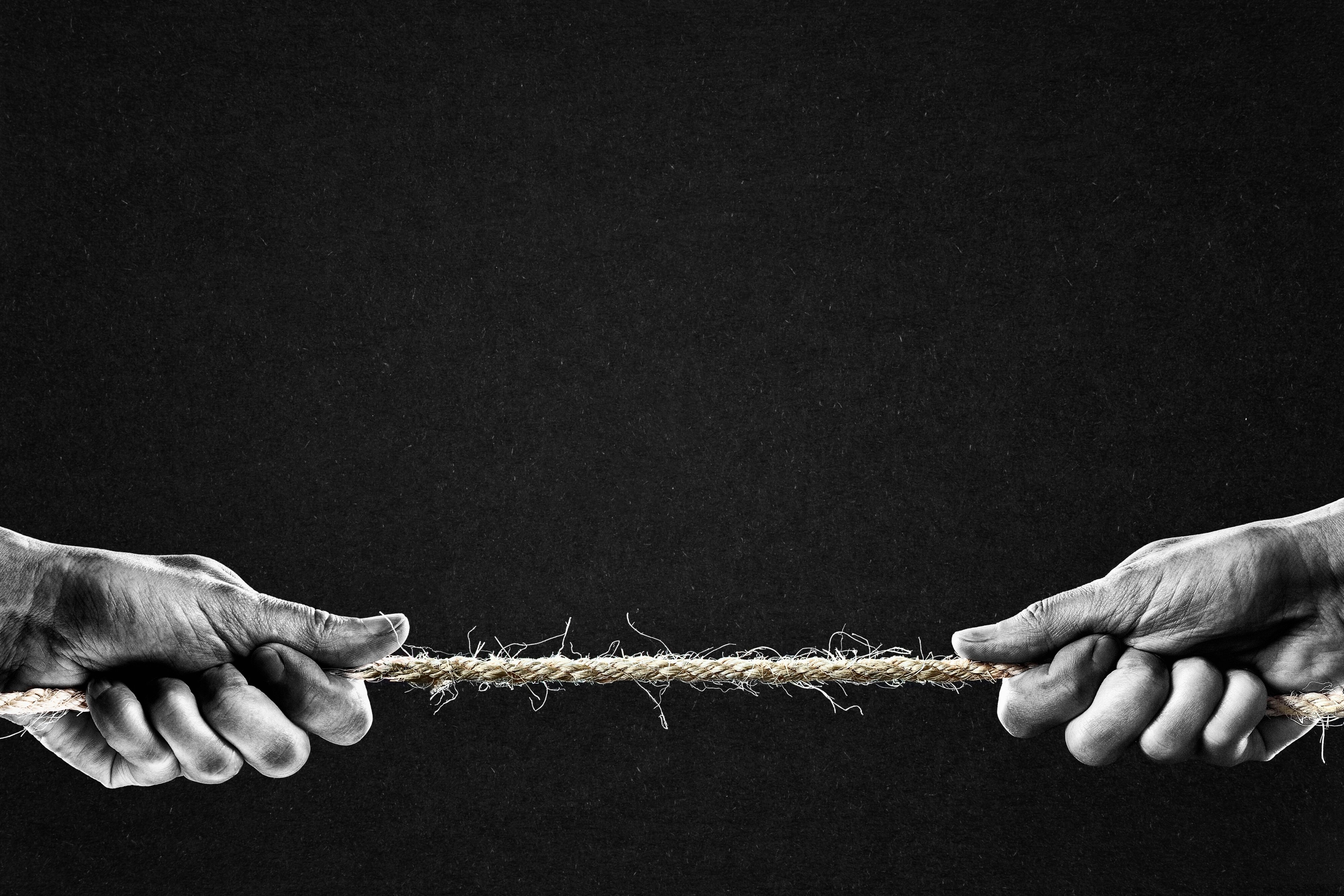 Two hands pulling on opposite ends of a frayed rope against a dark background, suggesting tension or struggle.