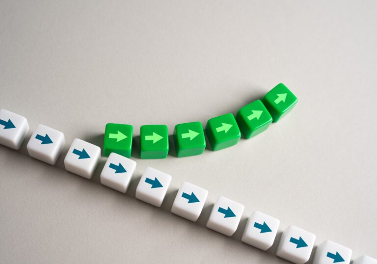 A row of white cubes with blue arrows moves in a straight line, while a series of green cubes with light green arrows curves upward, symbolizing innovation, change and reinvention.
