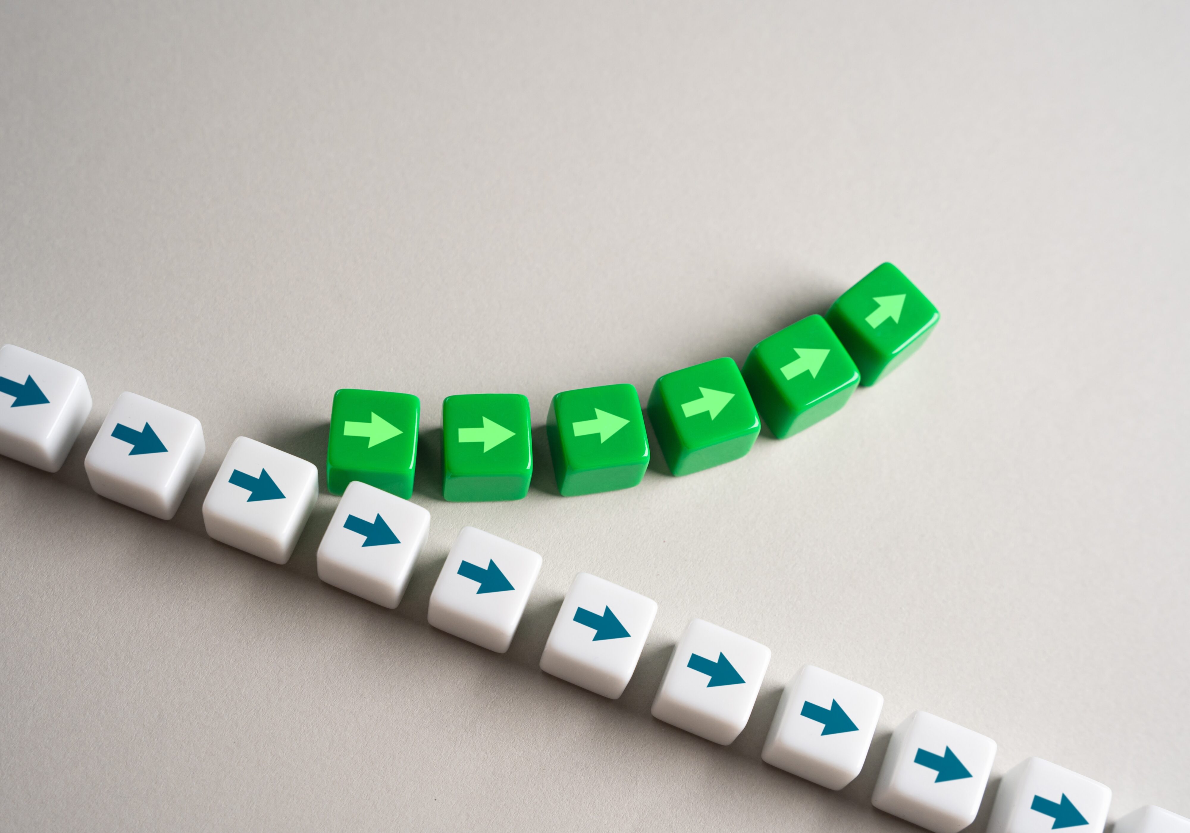A row of white cubes with blue arrows moves in a straight line, while a series of green cubes with light green arrows curves upward, symbolizing innovation, change and reinvention.