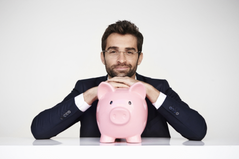 A man in a suit and glasses rests his hands on a large pink piggy bank while sitting at a white table, looking directly at the camera against a plain background.