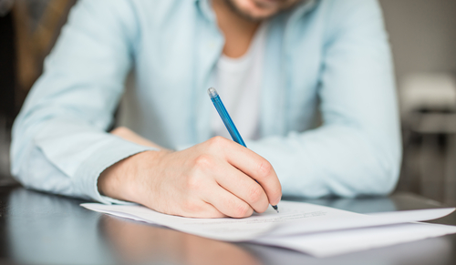 A person sits at a desk writing a letter with a blue pen, symbolizing the process of drafting an SBA loan hardship letter. Represents a business owner carefully explaining their financial situation and requesting relief from their lender.