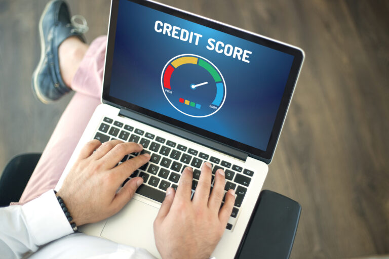 A person sits with a laptop on their lap, viewing a colorful gauge labeled “Credit Score” that ranges from poor to good. Their hands are on the keyboard, suggesting they’re checking or managing their financial standing online. The scene conveys focus, control and awareness—someone taking charge of their financial future.